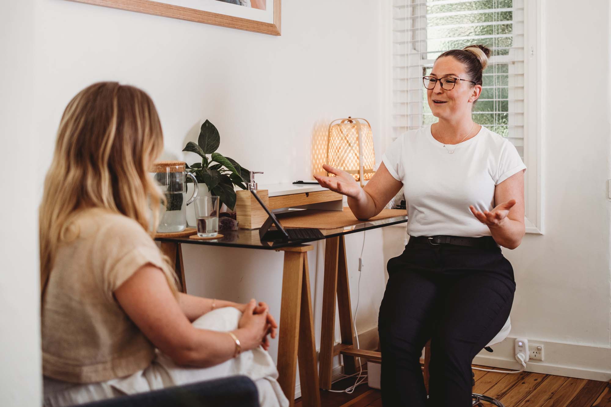 Two women are having a conversation in a home office at Akasha Integrative Health; one speaks while the other listens. A desk, water glasses, and a lamp are visible in the background.