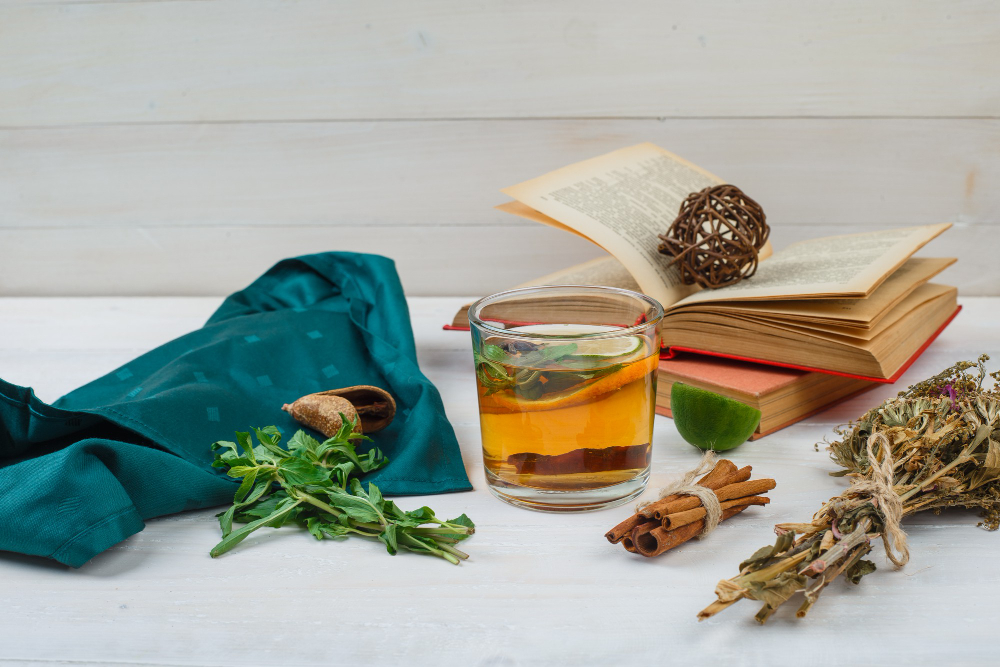 A glass of herbal tea sits on a white table surrounded by fresh herbs, cinnamon sticks, a green cloth, tied dried plants, and two open books—inviting quiet reflection on the philosophy of herbal medicine.