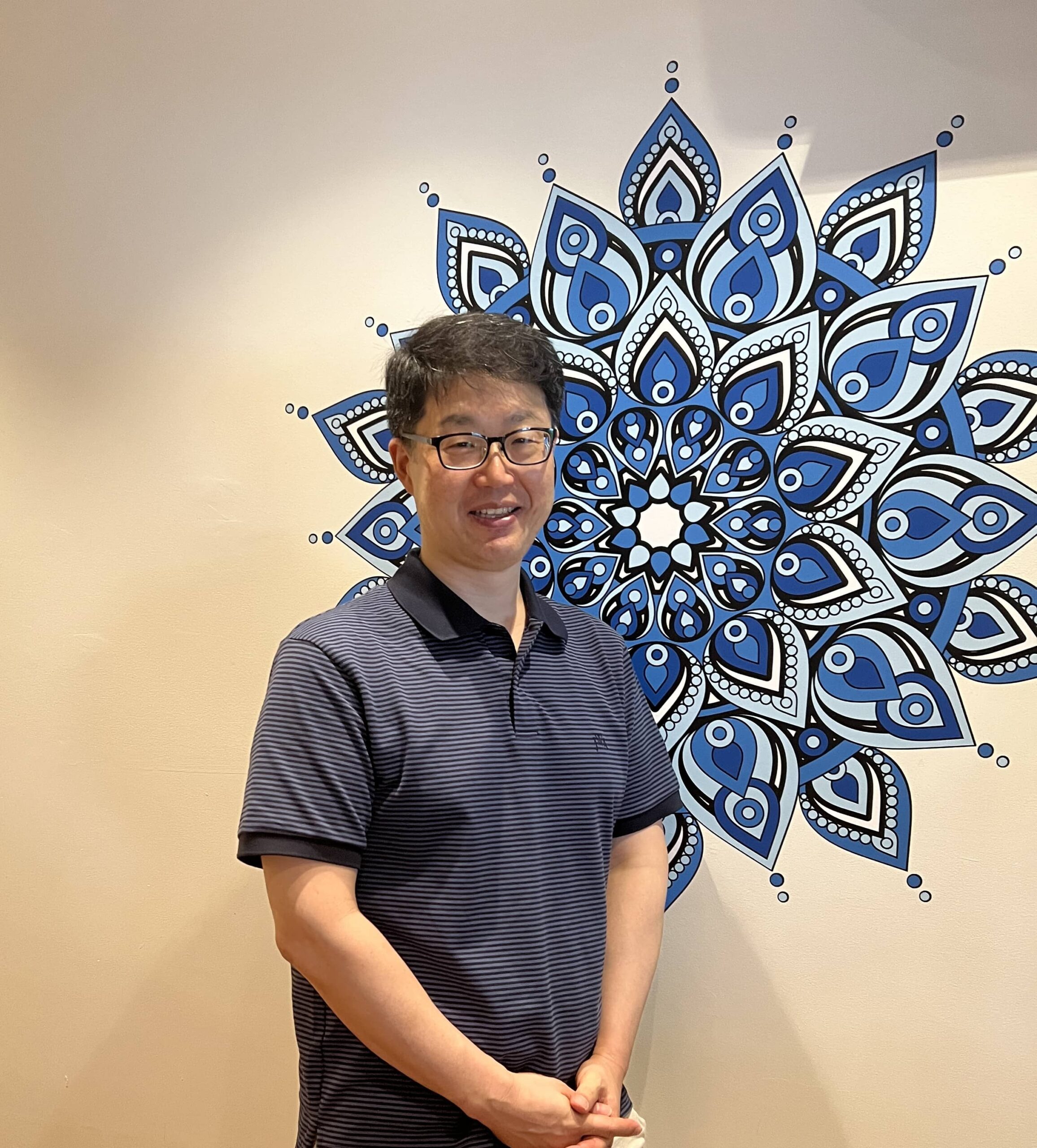 Charles Jwa, in glasses and a navy striped polo, stands before a large blue and white mandala design on the wall.