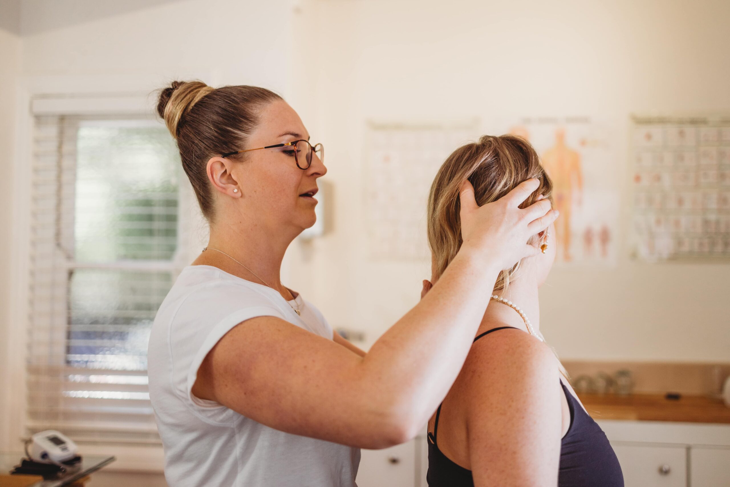Woman in glasses examines another woman's head and neck for Chiropractic Care; anatomical charts in background.