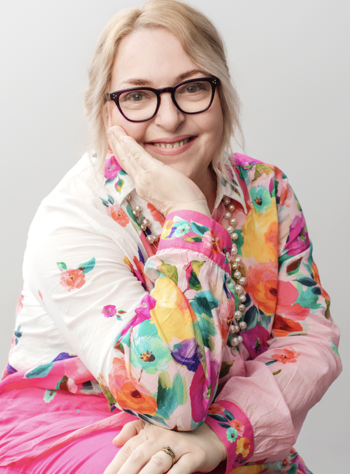 Marianne Edwards, blonde woman with glasses in a floral shirt and pearls, smiling with chin on her hand, gray background.