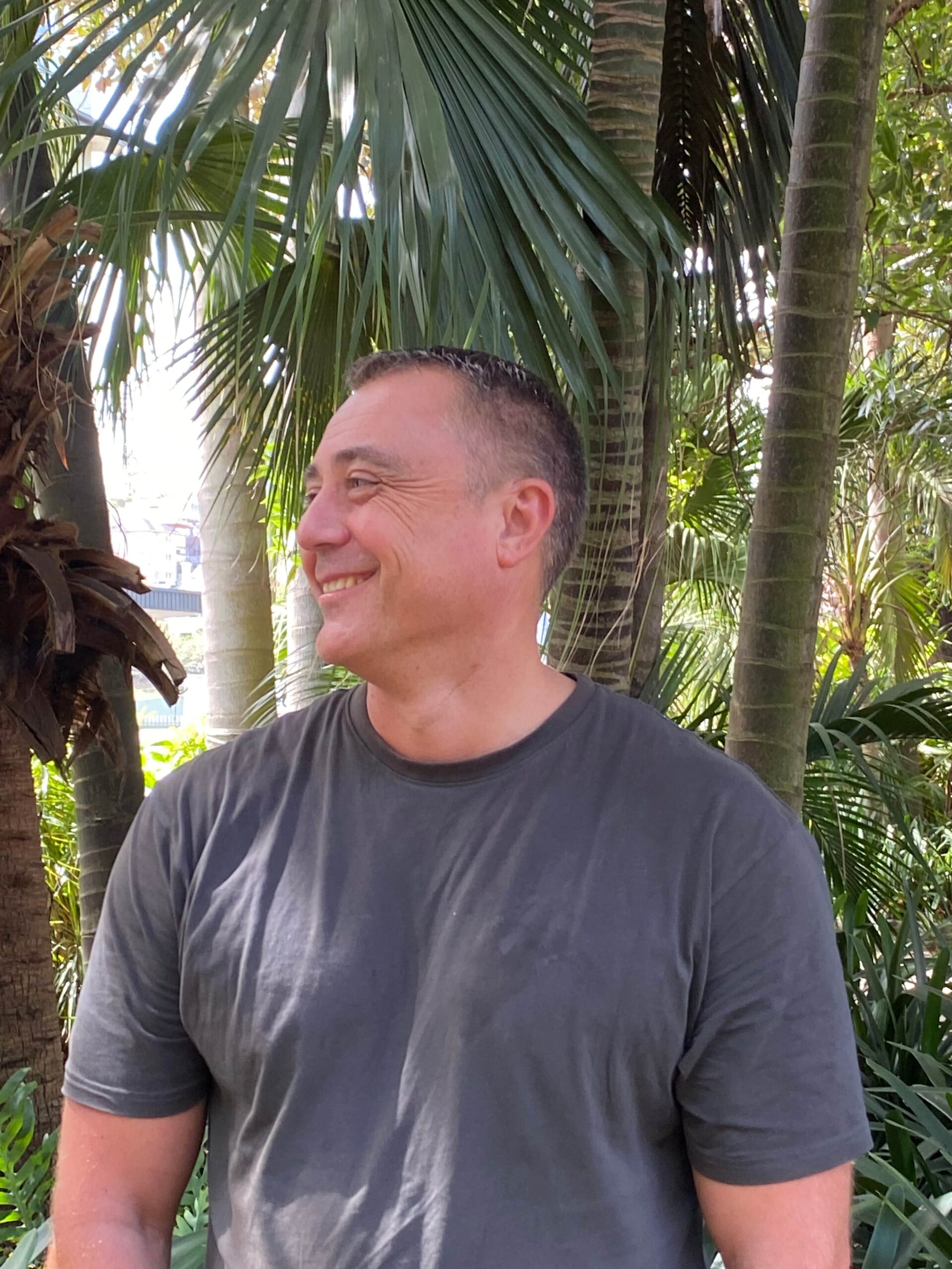 Phillip Mawson in a gray T-shirt smiles outdoors, looking to the side among tall green palm trees and lush foliage.