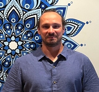 Dr Conna Alexander wears a blue shirt, standing before a wall with a bold blue and white geometric mandala pattern.