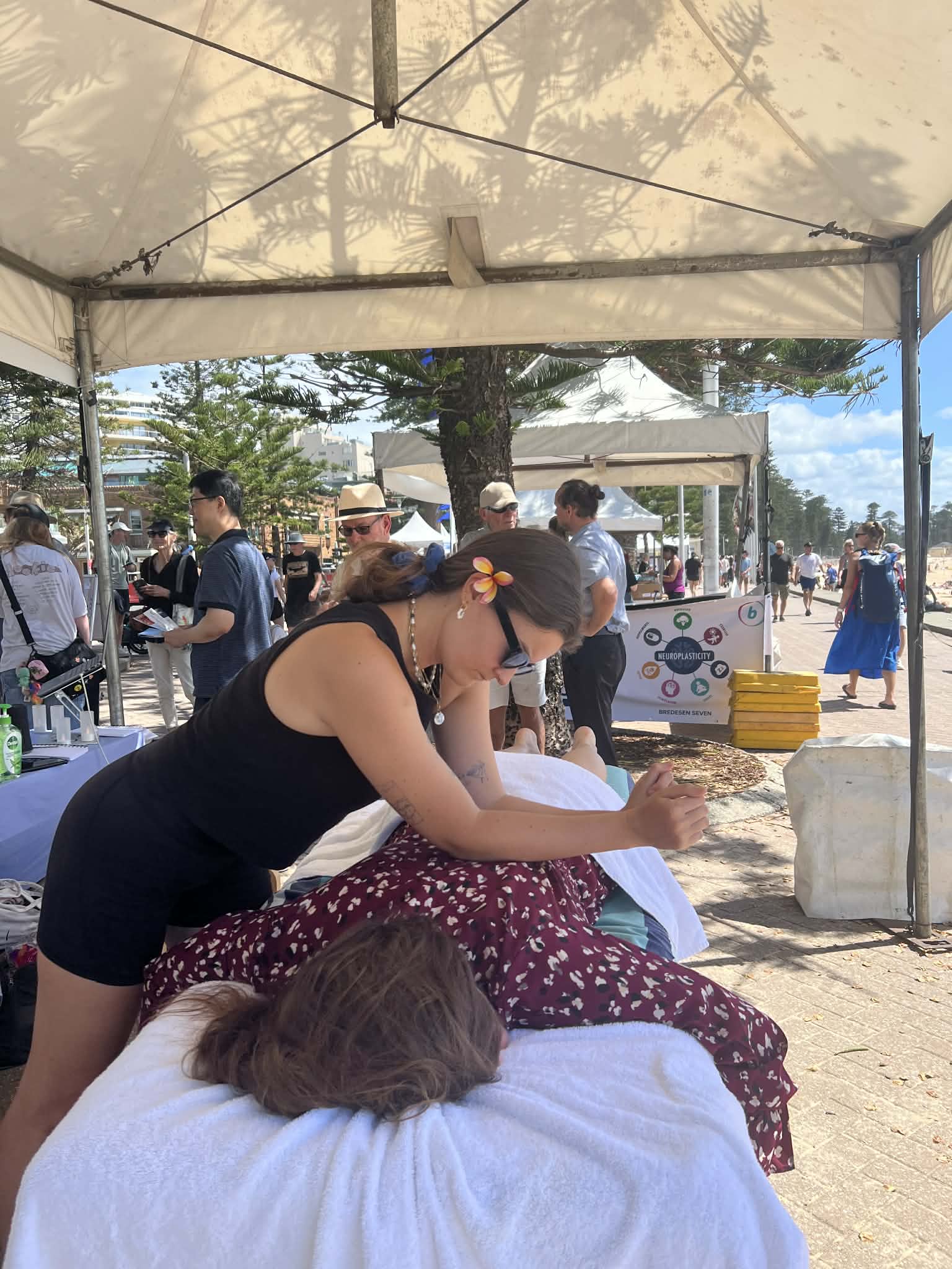 Remedial Massage at outdoor beach event—Akasha Health staff massages client under canopy, people relax nearby.