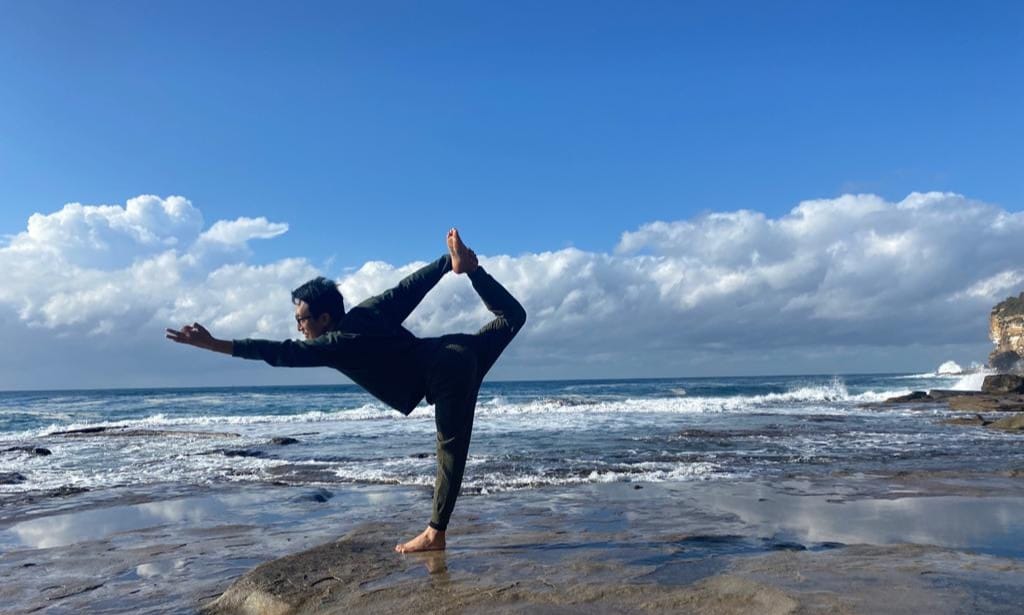 Person balancing in yoga pose on rocky shore, embracing Specialised Modalities with ocean and cloudy sky as backdrop.